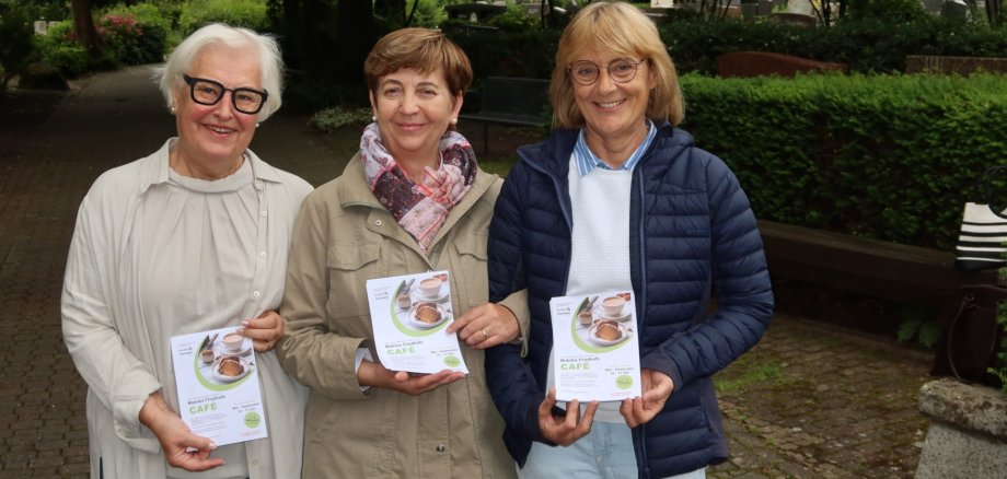 Drei Frauen stehen auf einem gepflasterten Weg in einem Park und halten jeweils ein Informationsblatt mit dem Titel 'Café' in den Händen.
