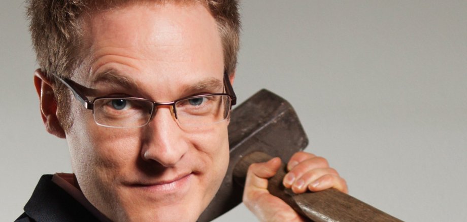 شخص ذو شعر أشقر قصير يرتدي بدلة سوداء وقميصاً وردياً فاتحاً، ويحمل فأساً ثقيلاً على كتفه