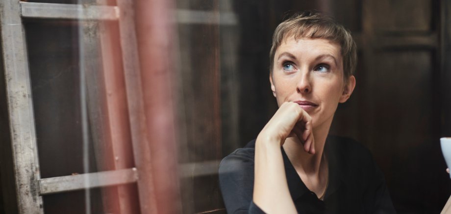 Човек с къса руса коса седи в тъмна стая. Облечено е в черен топ и държи чаша в дясната си ръка. На заден план се виждат дървена стълба и стена.