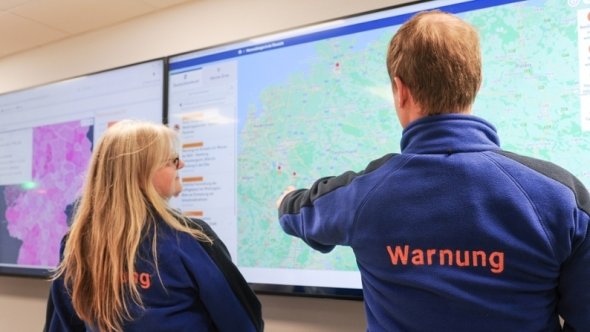 Two people in blue jackets bearing the word ‘Warning’ are looking at large screens displaying maps and data.