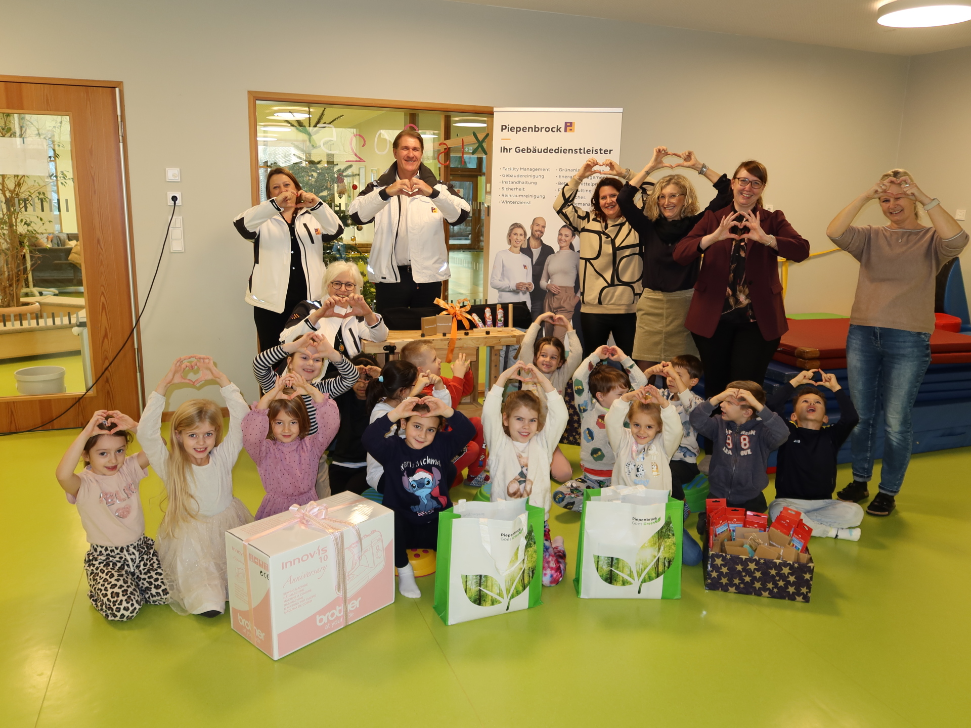 Group of adults and children at Kita 18, forming heart shapes with their hands, in front of several gift packages and a Piepenbrock banner.