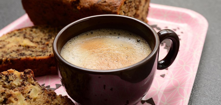 Schwarze Tasse mit Kaffee auf einem rosa Tablett, umgeben von zwei Scheiben Kuchen und einem ganzen Kuchenlaib.