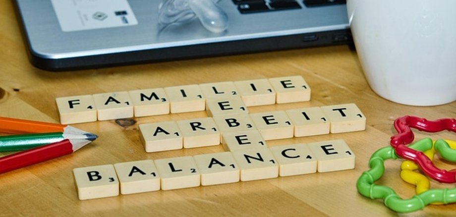 Scrabble tiles form the words 'FAMILY', 'WORK' and 'BALANCE' on a wooden table next to a laptop, a dummy, coloured pencils, a mug of coffee and colourful toys.