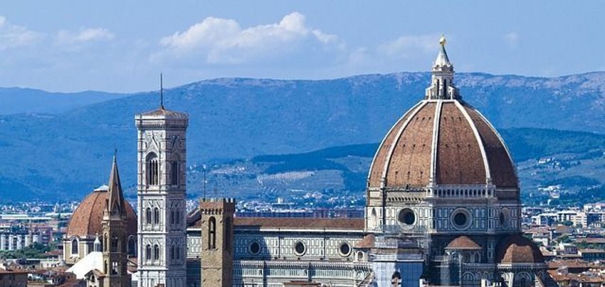 View of the dome and bell tower of Florence Cathedral against a backdrop of mountains and a city with red-tiled roofs