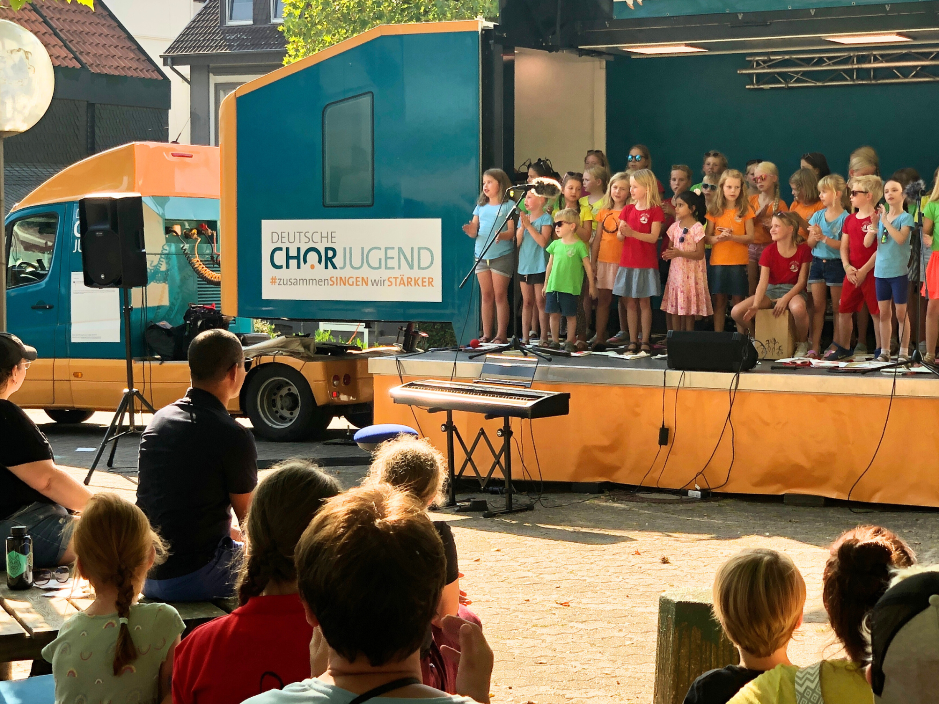 Children's choir sings on a stage in front of a blue truck with the lettering 'DEUTSCHE CHORJUGEND #zusammenSINGEN wir STÄRKER'