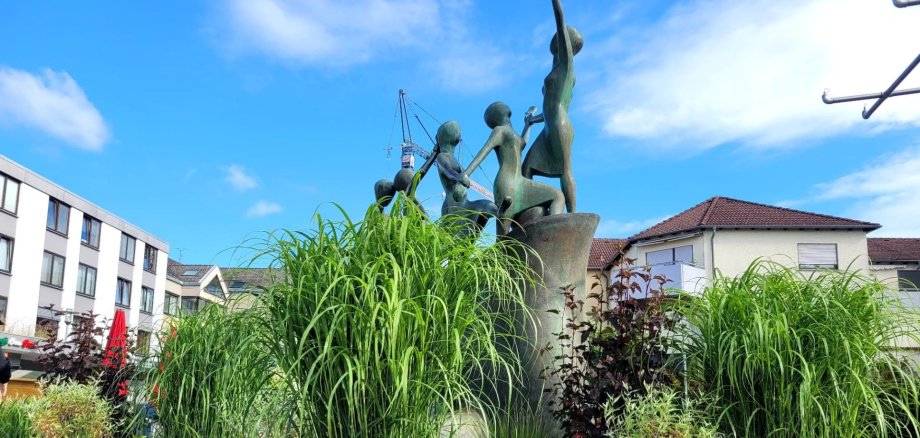 A fountain with greenery on Puiseaux Square A fountain on Puiseaux Square featuring a sculpture comprising several figures, surrounded by tall grasses and plants, with residential and commercial buildings in the background