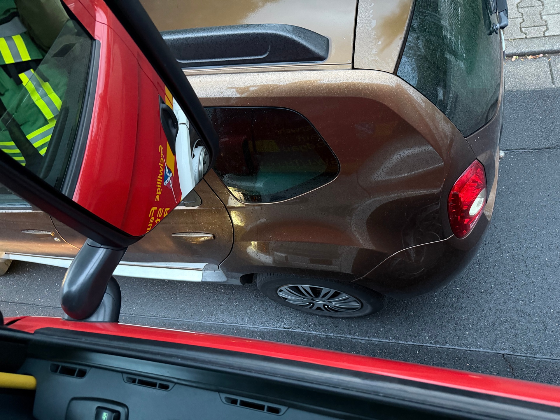 A view from above of a brown car parked on a road next to a red vehicle, with the driver wearing reflective fire service clothing