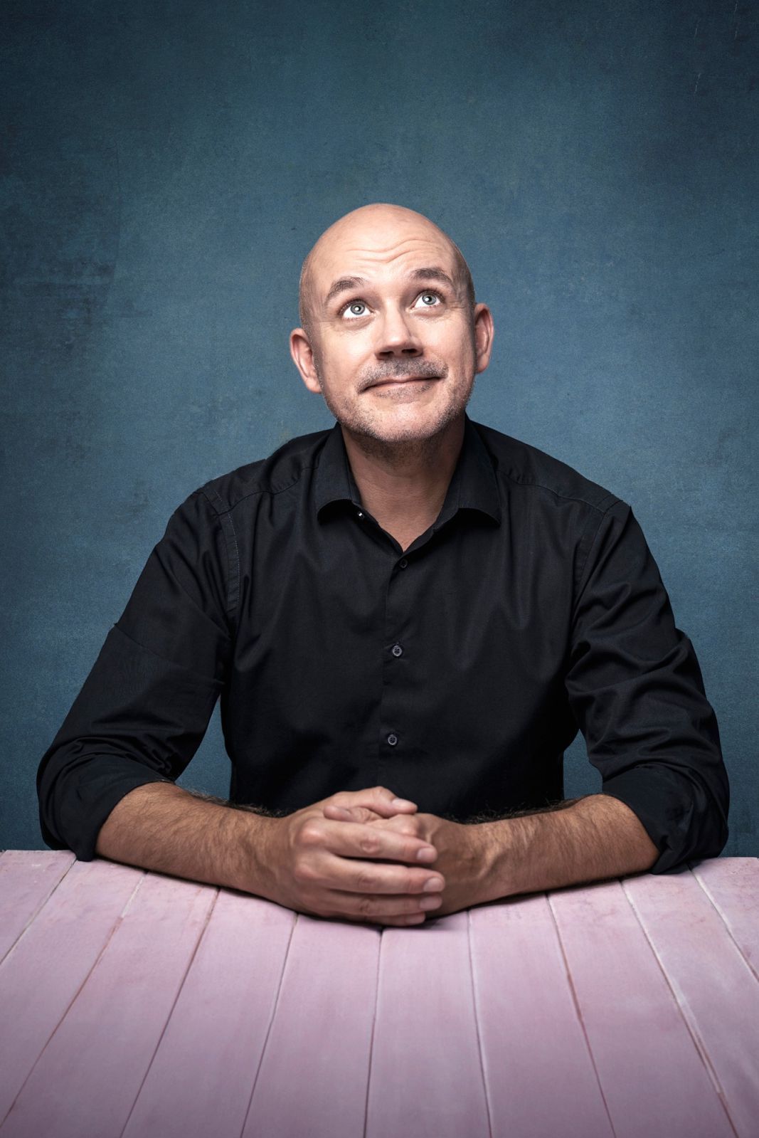 A bald man in a black shirt is sitting at a light-coloured table, looking up
