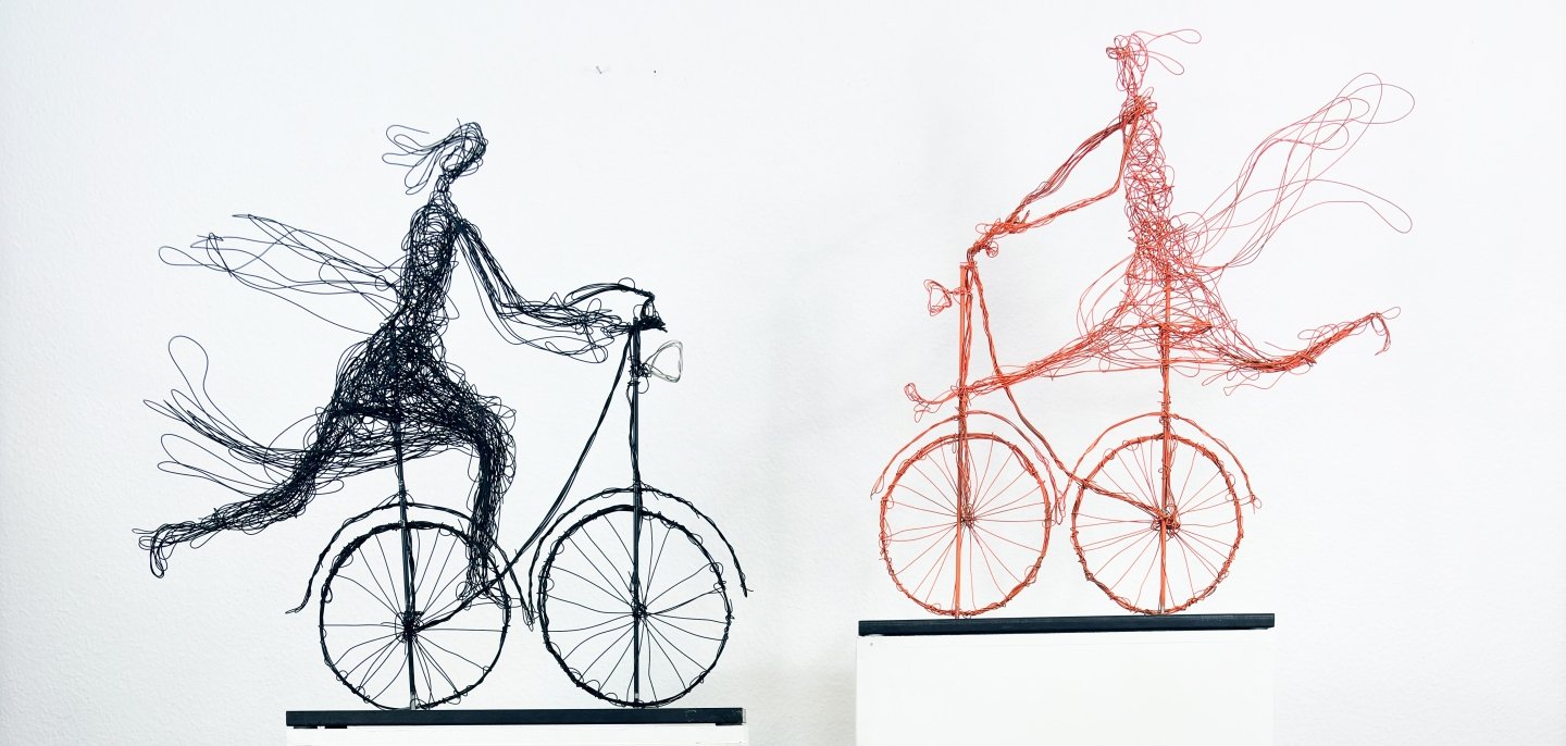 Artwork by Henriette Tomasi Two delicate wire sculptures of figures on bicycles, set on white plinths against a white wall