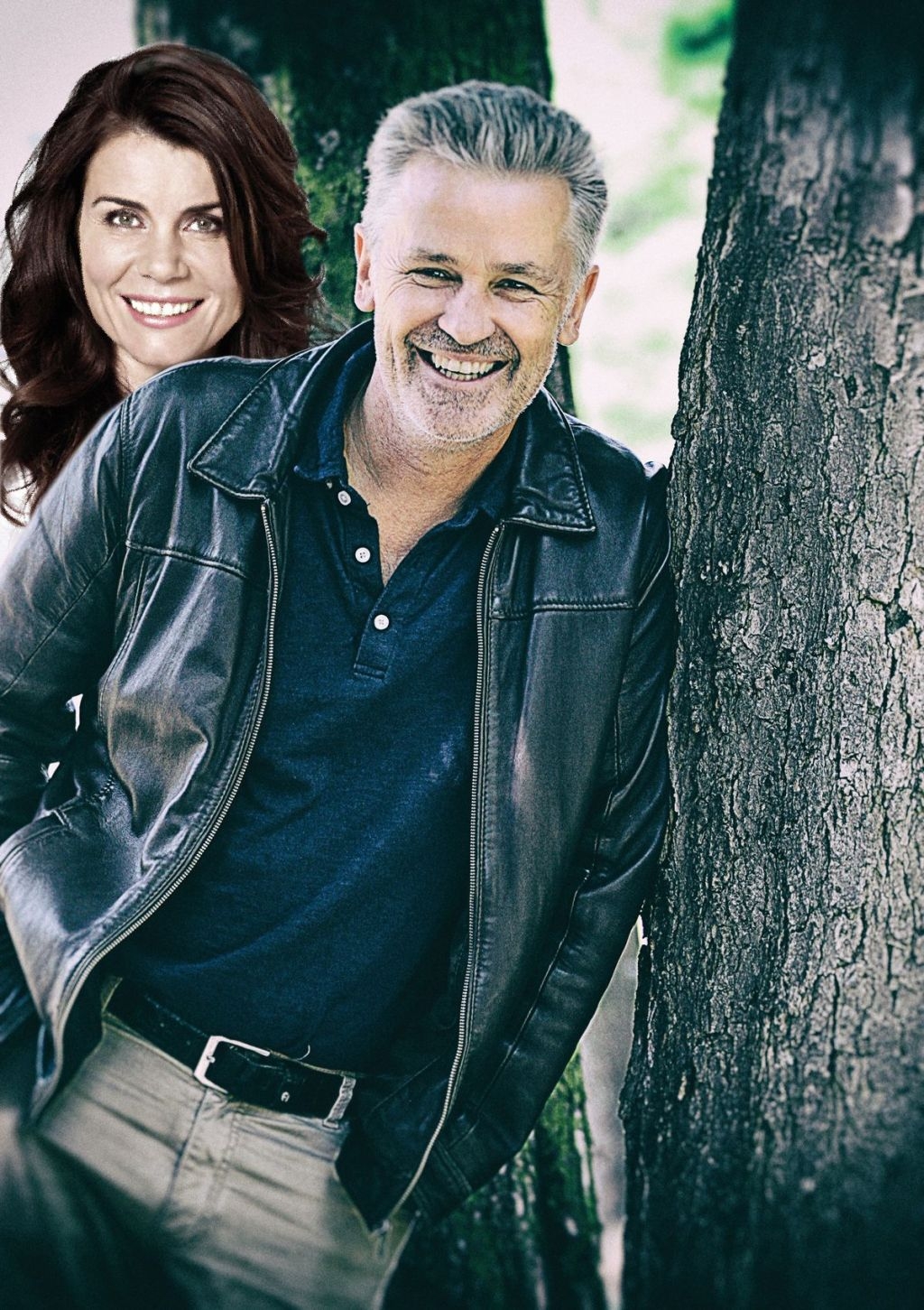 Two people are leaning against a tree, the man is wearing a black leather jacket and a dark shirt, the woman has long brown hair