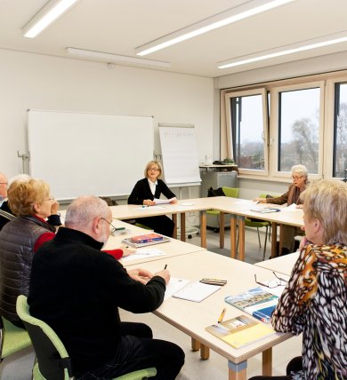 Training situation A group of older people are sitting at U-shaped tables in a bright training room with large windows, writing or listening