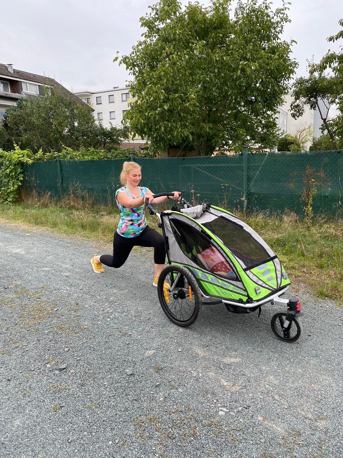 Woman in sportswear makes lunge with child trailer on gravel path outdoors