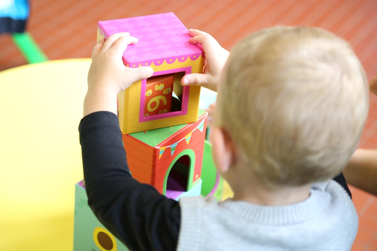 A toddler with blonde hair stacks colourful dice on a table. The cubes are designed in different colours and patterns.