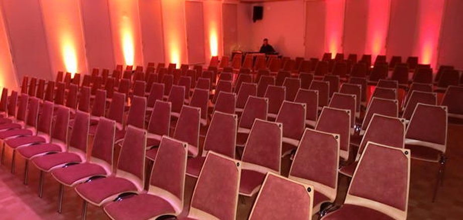 Hall in the Niederroden community centre with several rows of red chairs and orange-red wall lighting