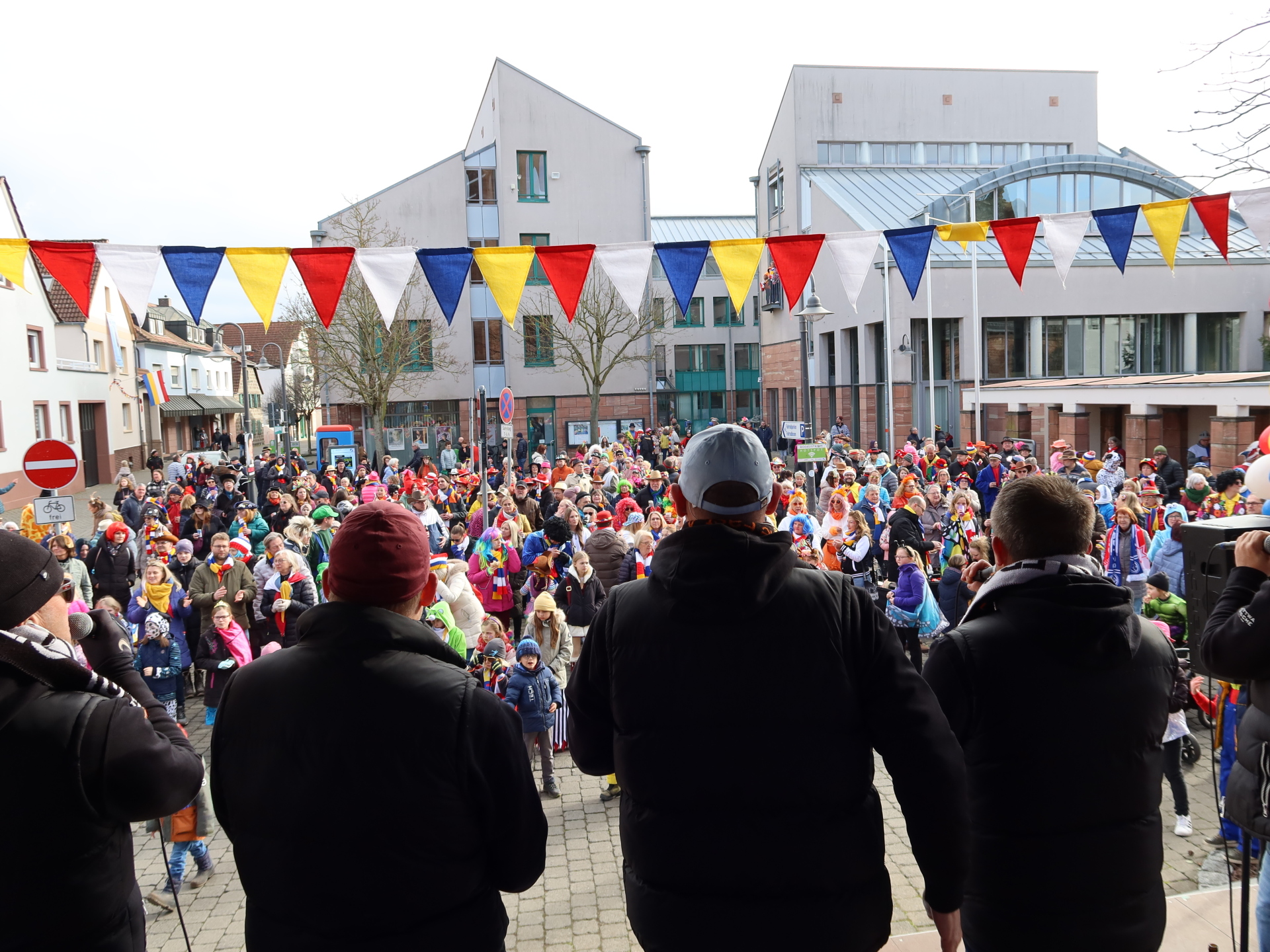 Crowd in front of the town hall during carnival, colourful pennants hanging over the scene. Three people with black jackets stand in the foreground, their backs to the camera.