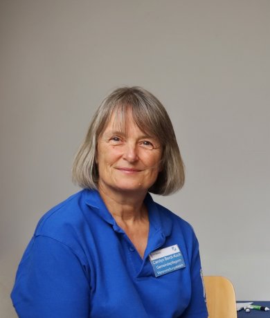 Parish nurse Mrs Berck-Koch Portrait of Berck-Koch in front of the town council meeting room. She has blonde-grey hair and is wearing a blue blouse.