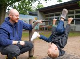 In the foreground, a child is doing gymnastics on a horizontal bar, with an educator squatting to the right. In the background is a day care centre.