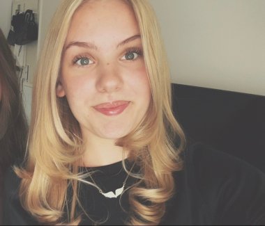 Sophia Rhein Portrait of a young woman with long blonde hair, slightly curled; she is wearing a black top and a necklace with a wing-shaped pendant