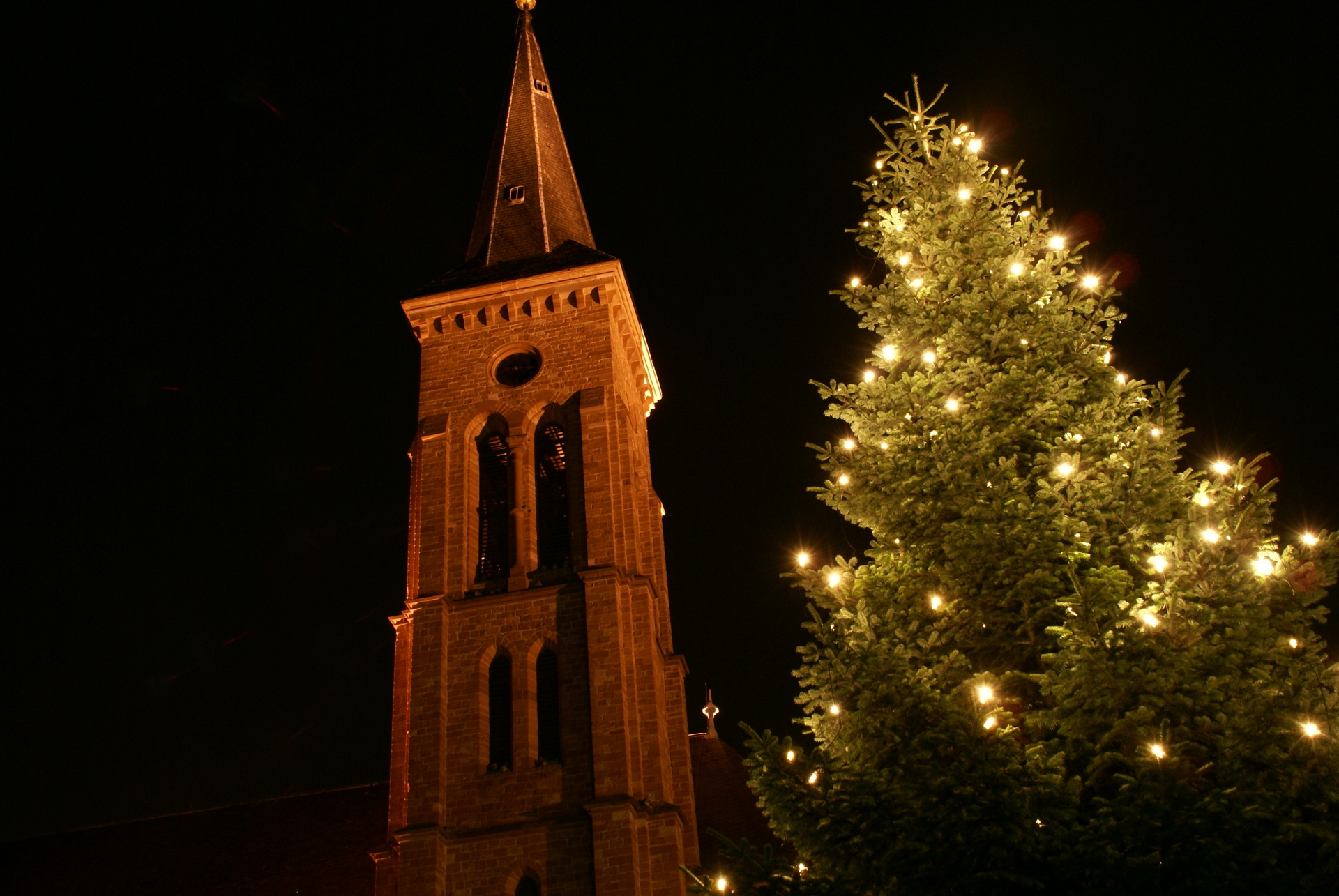 The illuminated church tower of St Nicholas' Church in Jügesheim next to a large Christmas tree decorated with lights at night.