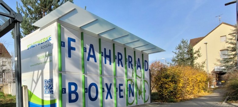 Several white bicycle boxes with blue and green lettering 'FAHRRAD BOXEN' at Dudenhofen railway station, surrounded by pavement and trees.