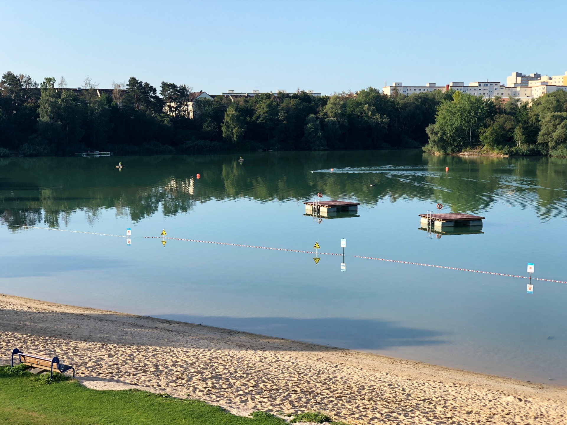 Vista del lido con dos plataformas flotantes. Playa de arena y pradera verde en la orilla. Árboles y edificios residenciales al fondo.