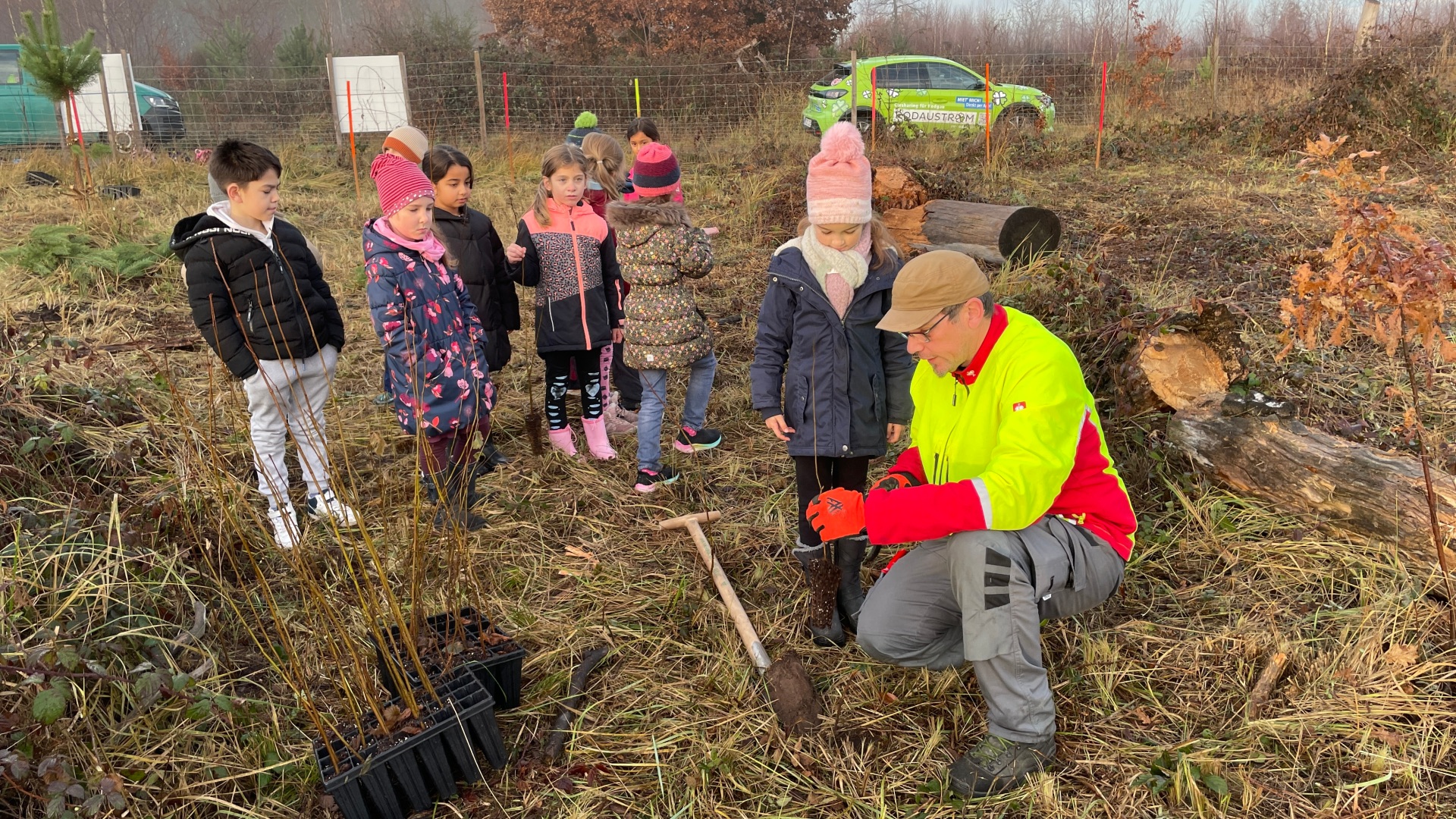 Grupo de niños y un empleado municipal con ropa de advertencia plantando árboles jóvenes en un campo con herramientas y plantones de árboles.