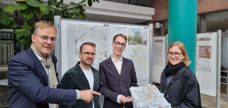 Cuatro personas, de izquierda a derecha Marcus Hille (presidente del jurado), Valentin Martin, Fabian Wieczorek y Pauline Rohländer (de ROHWAREIMSTUDIO) ante los planos arquitectónicos del diseño con el modelo ganador.