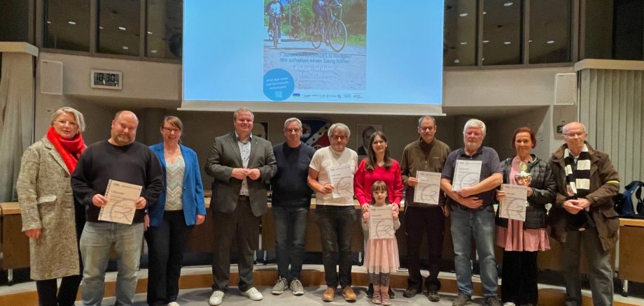 Grupo de doce personas de pie en un escenario, algunas sosteniendo certificados, al fondo una imagen de proyector con motivo de bicicleta y texto sobre el acto de clausura de CITY CYCLING.