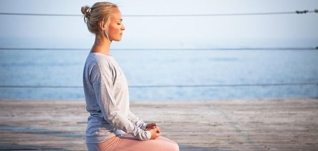 Una persona está sentada en posición de loto en una terraza de madera junto al agua. Lleva un top gris y unos leggings rosas. El fondo muestra aguas tranquilas y un cielo despejado.