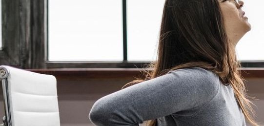 Dos mujeres en ropa deportiva. La mujer de la izquierda lleva una camiseta negra y unos leggings con un estampado morado. La mujer de la derecha lleva un top negro y unos leggings negros, con zapatos rosas. La mujer de la izquierda está apoyando a la mujer de la derecha, que está de pie en una pose deportiva.