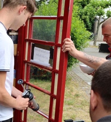 Acción 16 Plus Un joven sujeta una puerta de madera roja mientras otra persona con un brazo tatuado estabiliza la puerta y una tercera sujeta un smartphone en un trípode