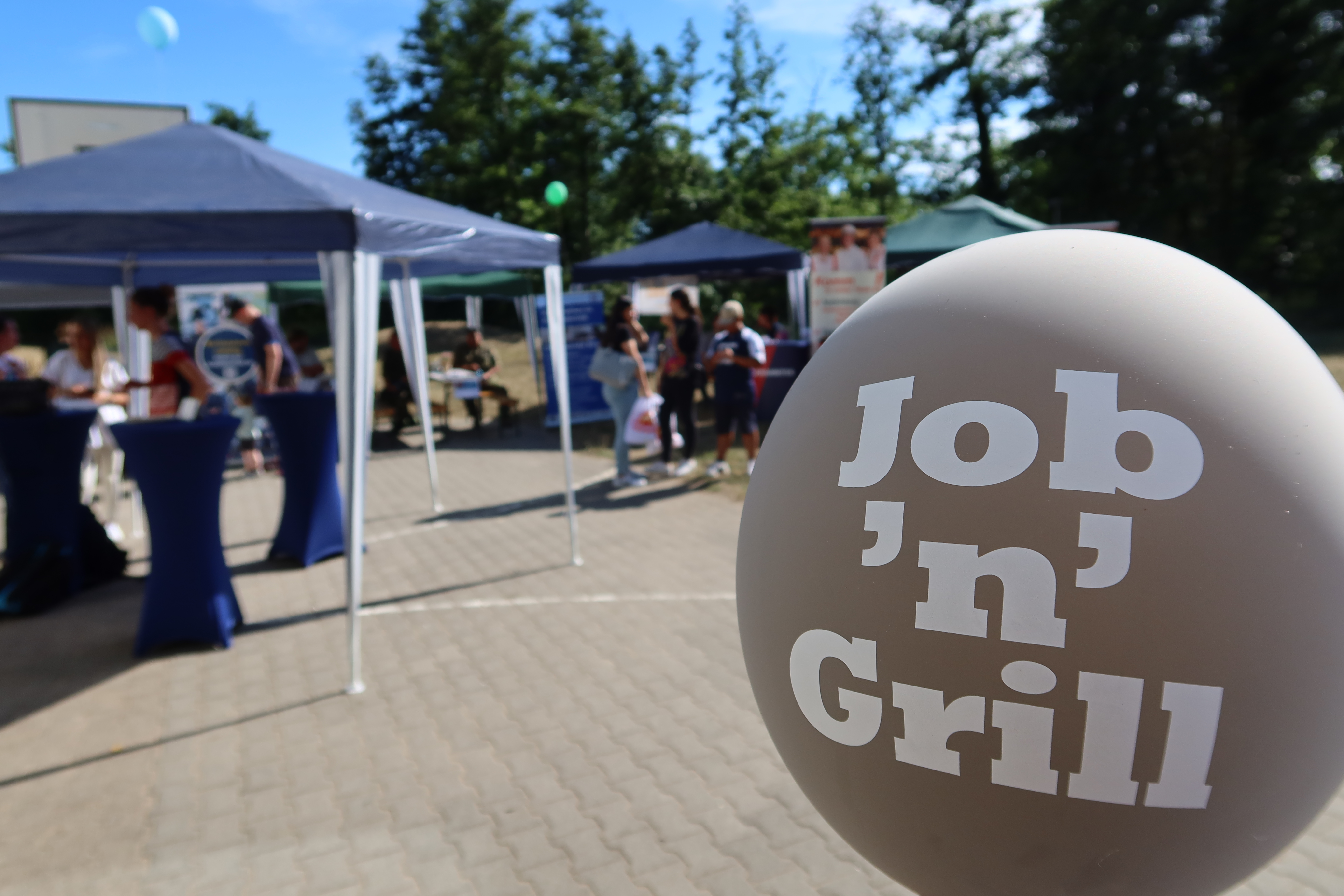 Globo con la etiqueta "Job 'n' Grill" ante los stands y visitantes de una feria de educación al aire libre