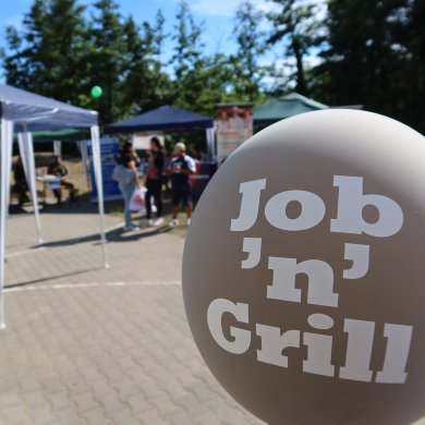 Feria de la educación Globo con la etiqueta "Job 'n' Grill" ante los stands y visitantes de una feria de educación al aire libre