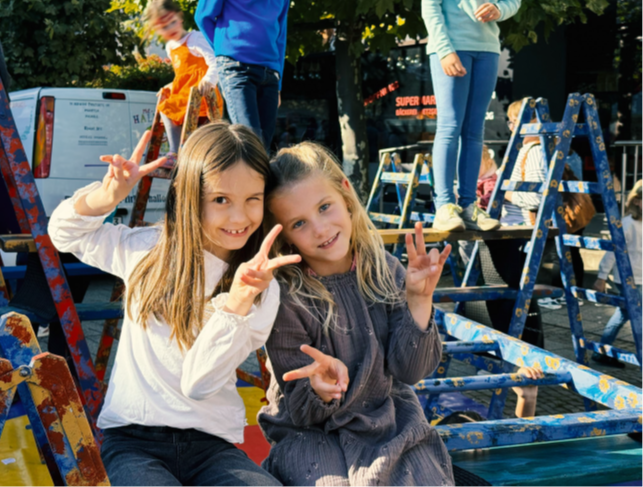 Dos niñas levantan las manos con el signo de la victoria. Al fondo, unos niños suben por un recorrido de escaleras.