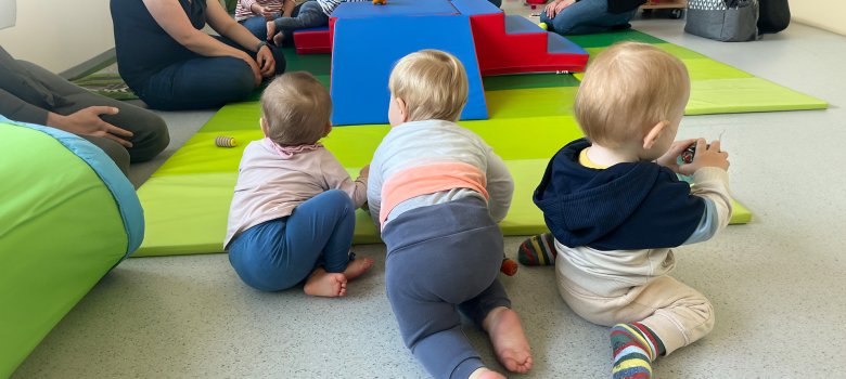 Tres niños pequeños Tres niños pequeños están sentados y gatean sobre una alfombra de color verde claro. Al fondo se ve a otros niños y adultos sentados alrededor de una estructura de juegos azul y roja. Los niños llevan ropa de diferentes colores, entre la que destacan calcetines y jerséis a rayas. Hay juguetes esparcidos por la alfombra.