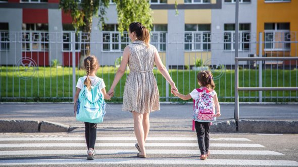 Mujer con dos niñas Una mujer coge de la mano a dos niñas con mochilas mientras cruzan un paso de cebra frente a un colorido edificio residencial