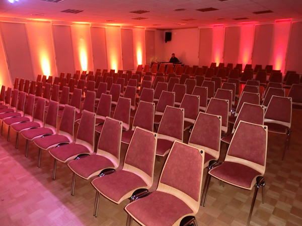 Sala del centro comunitario de Niederroden con varias hileras de sillas rojas e iluminación de pared en rojo anaranjado