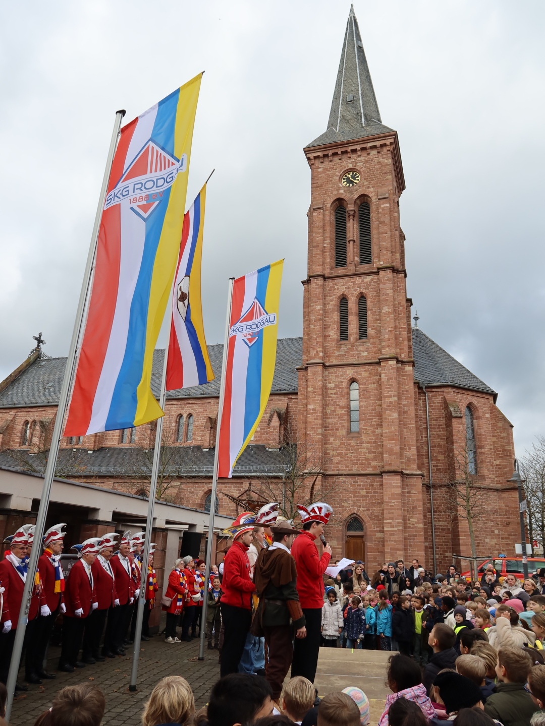 Las banderas del carnaval están izadas, los funcionarios de pie en el escenario, rodeados por el público al fondo está la iglesia de San Nicolás.