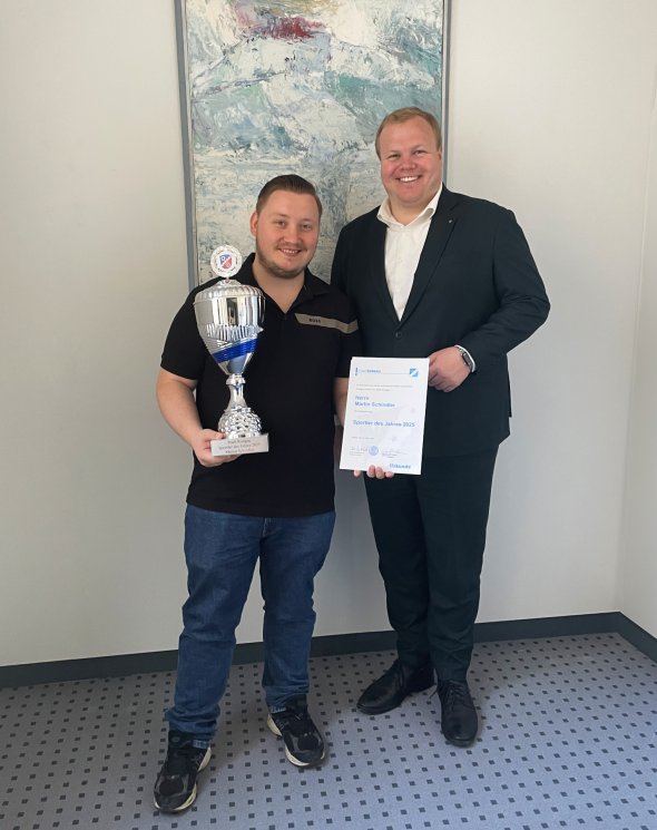 Martin Schindler - Deportista del año 2025 en Rodgau Martin Schindler y el alcalde Breitenbach de pie uno junto al otro con un cuadro abstracto en la pared, Schindler a la izquierda sosteniendo un gran trofeo, el alcalde a la derecha un certificado