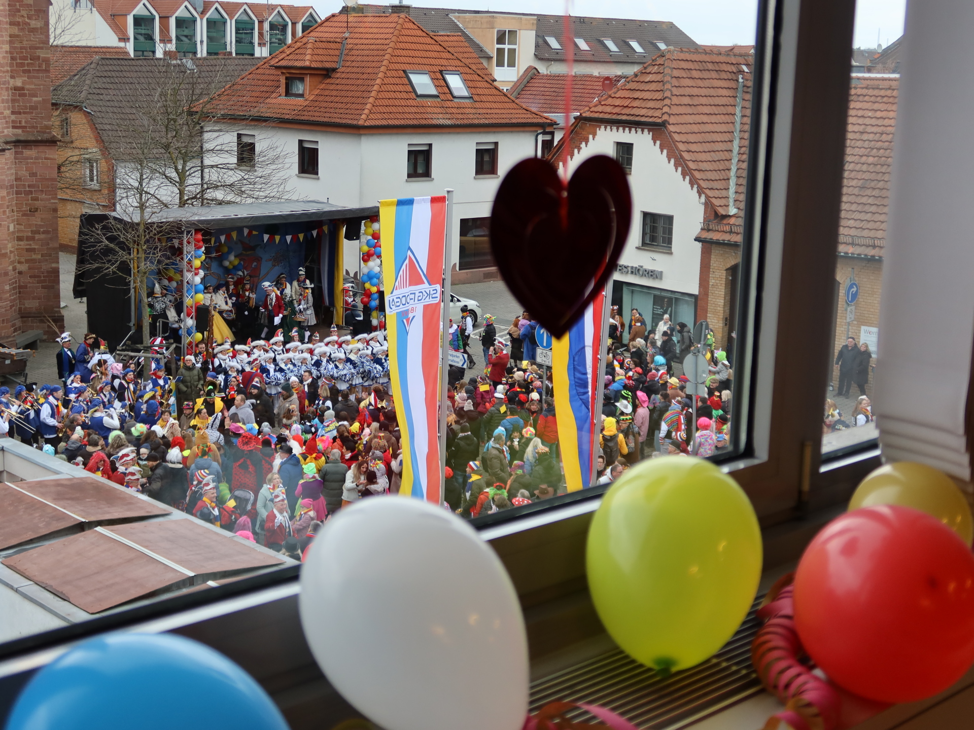 Vista desde una ventana de una multitud colorida frente a un escenario con banderas y globos en primer plano