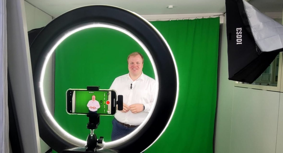 El alcalde Max Breitenbach, con camisa blanca y pantalones oscuros, de pie ante un fondo verde, fotografiado con luz anular con un smartphone sobre un trípode, rodeado de iluminación de estudio.