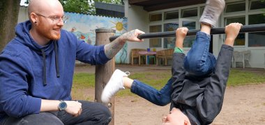 Prácticas reconocidas como educador En primer plano, un niño hace gimnasia en una barra horizontal, con un educador en cuclillas a la derecha. Al fondo se ve una guardería.