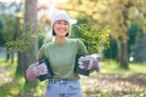 Una mujer vestida con ropa de jardinería sostiene plantas en macetas. Está de pie en un parque con árboles al fondo.