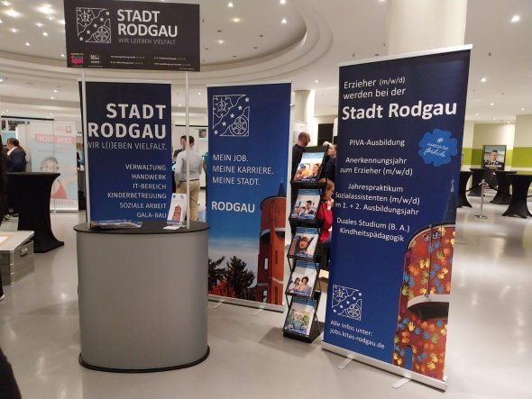 Stand de exposición azul de la ciudad de Rodgau con publicidad de carreras de educación. Junto a él se exhiben folletos. Al fondo se ven otros expositores de la feria.