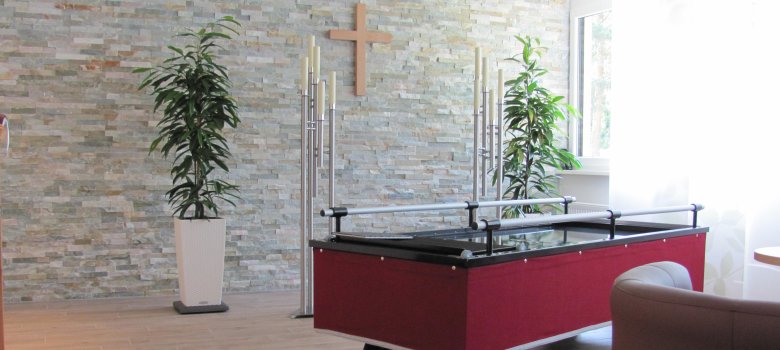 Habitación de despedida con carrito Al fondo hay un muro de piedra con una cruz, delante de la cual hay un carro en el que se puede depositar un ataúd.