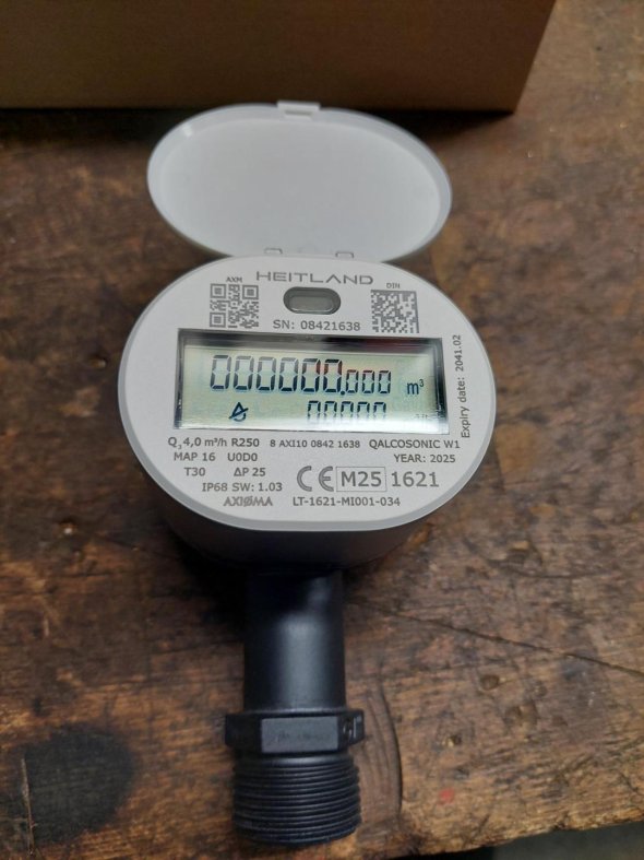 Un contador de agua por radio descansa sobre una base de madera con una pantalla abierta que muestra una serie de cifras y datos técnicos.