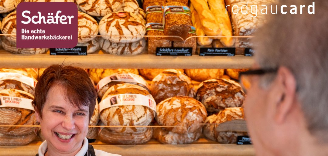 Boulangerie avec différentes sortes de pain en arrière-plan. Une personne en chemise rayée et tablier se tient derrière le comptoir. Un client vêtu d'une veste orange tient un smartphone en direction de la vendeuse. Texte : 'Information importante !