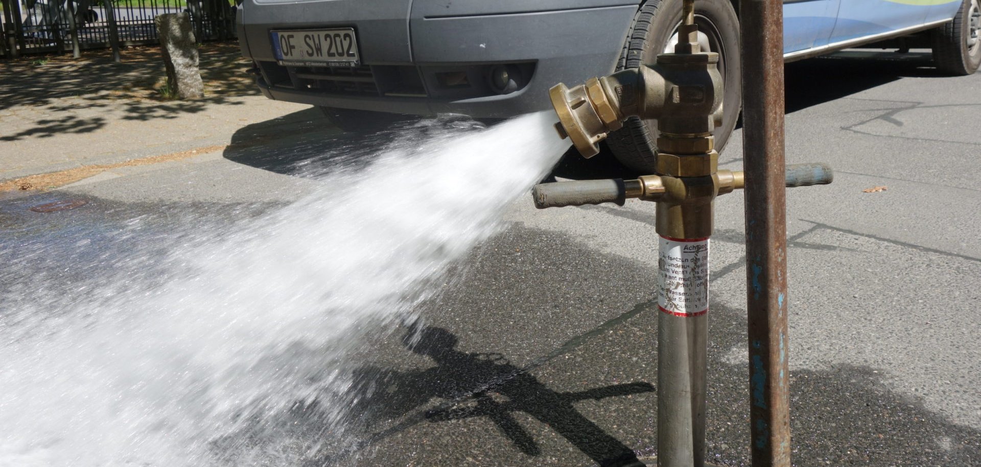 Un jet d'eau jaillit d'une bouche d'incendie sur le trottoir. En arrière-plan se trouve une camionnette blanche avec des marques vertes et rouges.