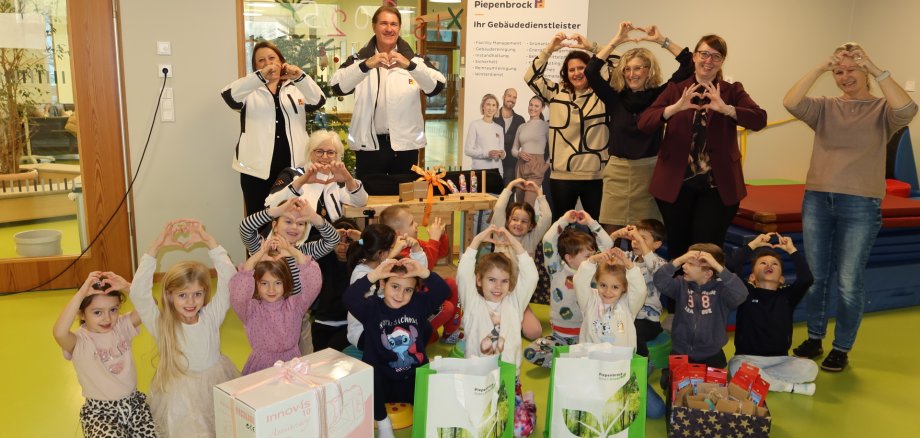 Groupe d'adultes et d'enfants à la Kita 18, formant des formes de cœur avec leurs mains, devant plusieurs paquets cadeaux et une bannière Piepenbrock.