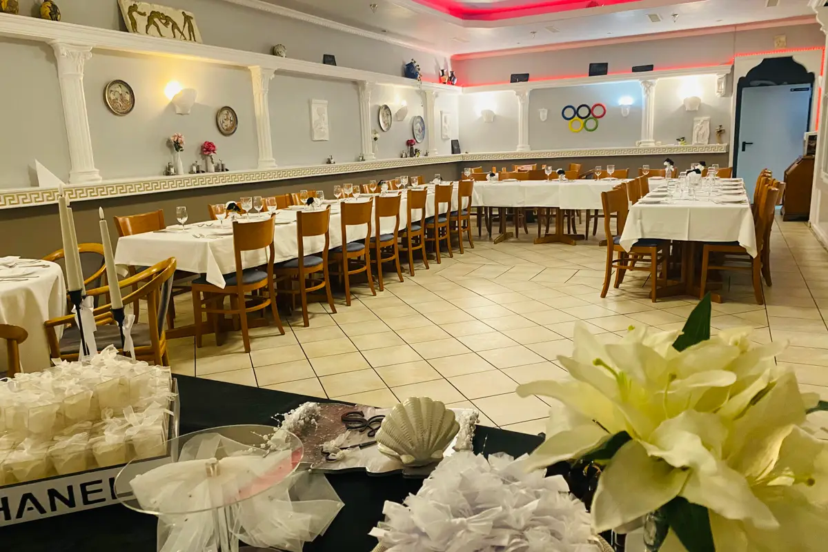 Grande salle de restaurant festive avec plusieurs longues tables, sol en bois et nappes blanches, décorée de fleurs et d'horloges murales.